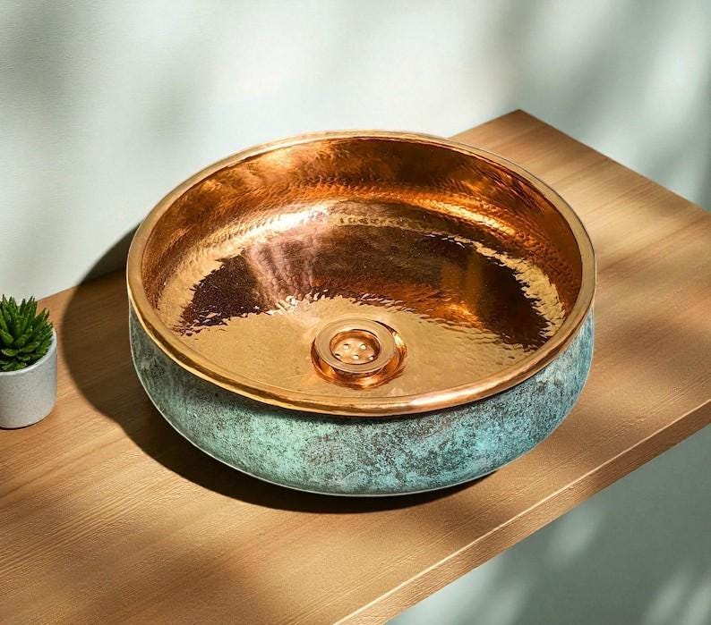 May include: A copper-colored, round vessel sink with a hammered interior and a verdigris exterior. The sink has a polished copper rim and a central drain. The sink is displayed on a wooden surface, showcasing its metallic sheen and unique color combination.