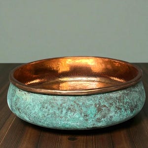 May include: A handcrafted copper bowl with a unique patina. The interior is a bright copper color, while the exterior has a turquoise-green finish. The bowl has a wide, shallow design, perfect for decorative use or serving.