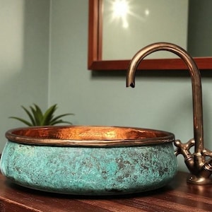 May include: A copper sink with a verdigris finish and a bronze faucet. The round sink sits on a dark wood surface. The faucet has cross handles. A small plant is visible in the background.