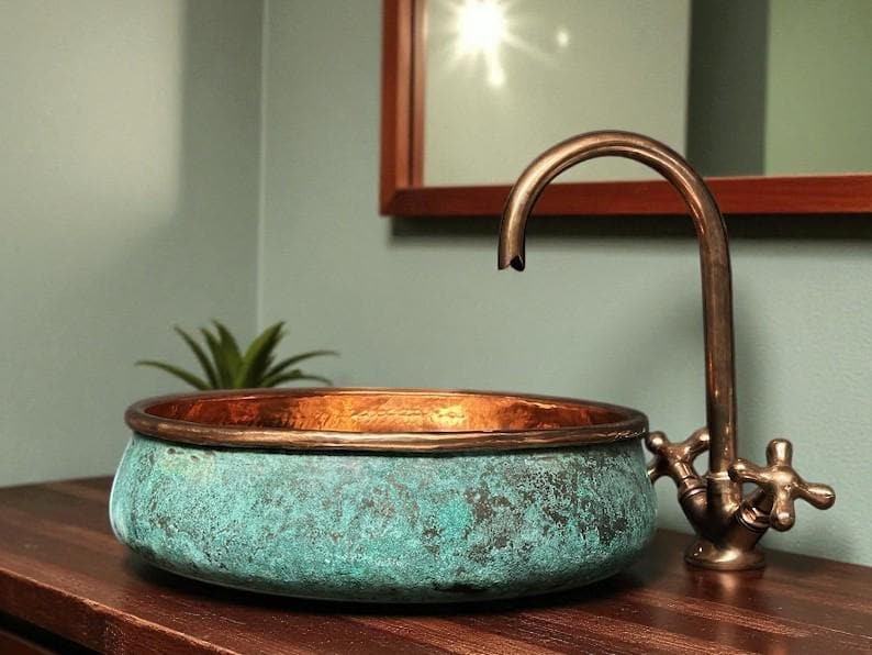 May include: A copper sink with a verdigris finish and a bronze faucet. The round sink sits on a dark wood surface. The faucet has cross handles. A small plant is visible in the background.