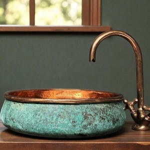 May include: A copper sink with a verdigris finish and a copper rim. The faucet is made of copper and has cross handles. The sink is on a wooden surface. A small potted plant is to the right.