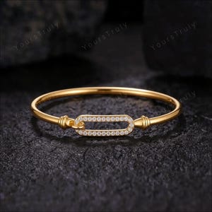 May include: A gold-toned bangle bracelet featuring a central horizontal oval link. The oval link is encrusted with small, clear, sparkling stones along its entire perimeter. The bangle has a smooth, polished finish and a simple, clean design.