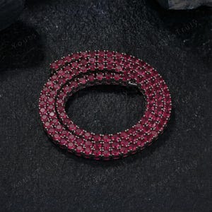 May include: A tennis chain necklace with numerous round, deep red gemstones set closely together. The necklace is displayed on a dark, textured surface, creating a contrast that highlights the vibrant color of the stones. The jewelry is a statement piece.