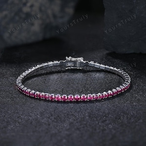 Könnte beinhalten: Ein silbernes Armband mit einer Reihe kleiner, leuchtend rosa Edelsteine. Das Armband hat einen sicheren Verschluss und wird auf einer dunklen, strukturierten Oberfläche präsentiert. Die Worte "Yours Truly" sind im Hintergrund schwach sichtbar.