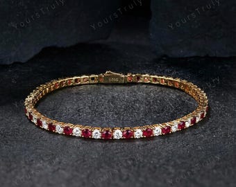 May include: A gold-toned bracelet with alternating red and white gemstones. The bracelet is set against a dark, textured background. The clasp is visible, and the inscription "S925" is present.