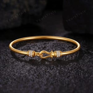 May include: A gold bangle bracelet featuring a central hook-and-eye style clasp mechanism. The two cylindrical sections flanking the clasp are encrusted with small, clear, round-cut stones, providing a contrast to the smooth, polished gold band.