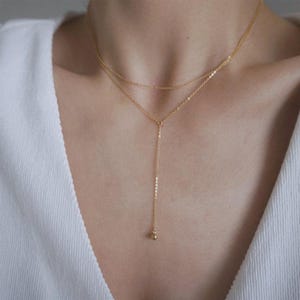May include: A delicate gold-colored necklace with two strands. The top strand sits close to the neck, while the second strand descends into a vertical chain ending with a small, round charm. The necklace is displayed against a white background.