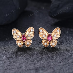 Puede incluir: Un par de pendientes de botón con forma de mariposa en oro. Cada pendiente presenta una piedra roja central de talla marquesa rodeada por cuatro pequeñas piedras blancas redondas incrustadas en las alas con textura veteada.