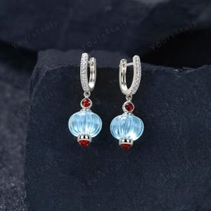 May include: A pair of silver hoop earrings featuring a dangling light blue ribbed glass bead shaped like a lantern. Each earring is accented with two small round red stones, one above and one below the glass bead, and clear pavé stones.