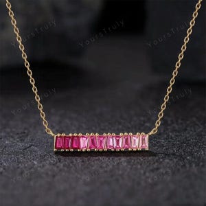 May include: A gold-colored necklace with a bar pendant featuring a gradient of pink rectangular gemstones. The chain is delicate, and the pendant is centered. The background is dark.