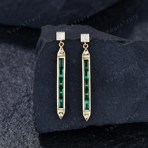 May include: A pair of gold drop earrings featuring a vertical bar design. Each earring contains four rectangular green gemstones set in a row, accented by a square white gemstone at the top and a small round white gemstone at the pointed base.