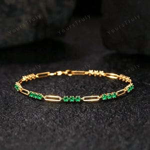 May include: A gold chain bracelet featuring a series of elongated oval links interspersed with small, round, vibrant green gemstones. The bracelet is constructed with a repeating pattern of metal links and clusters of three green stones.