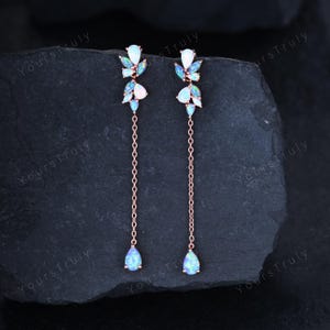 May include: A pair of rose gold dangle earrings featuring a cluster of marquise-cut opals at the top, connected to a delicate chain that ends in a single pear-shaped opal drop. The earrings exhibit iridescent flashes of blue and green color.