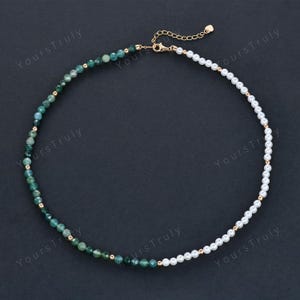 May include: A necklace featuring a split design with green gemstone beads on one side and white pearl beads on the other. Gold-colored beads and a gold clasp complete the necklace. The necklace is displayed on a dark background.