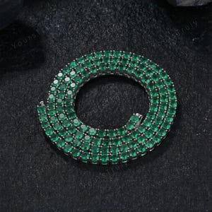 May include: A silver-toned necklace with a continuous row of emerald-green gemstones. The gemstones are round-cut and set closely together, creating a vibrant, eye-catching piece of jewelry. The necklace is displayed on a dark surface.