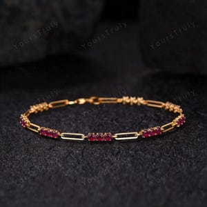 May include: A gold-colored bracelet with rectangular links and small, round, red gemstones. The bracelet is displayed on a dark, textured surface. The text "Yours Truly" is visible in the background.