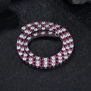 May include: A silver necklace with alternating rows of white and ruby-colored gemstones. The necklace is circular and rests on a dark, textured surface. The gemstones are round and evenly spaced.