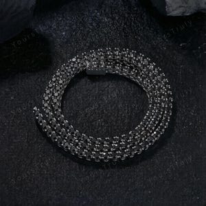 May include: A black tennis chain necklace with multiple rows of black stones. The necklace is displayed on a dark surface, showcasing its sparkling appearance. The clasp is visible, indicating the necklace's design and construction.