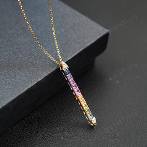 Op de afbeelding: Een gouden ketting met een verticale staafhanger bezet met een rij vierkant geslepen edelstenen in een kleurverloop van blauw, paars, roze, oranje, geel en wit, hangend aan een fijne gouden schakelketting.