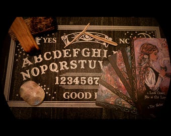 May include: A dark wooden Ouija board with white lettering and numbers, accompanied by tarot cards. A wooden planchette, a crystal, and burning sage create a mystical and spiritual setting. The board reads 'YES' and 'GOOD'.