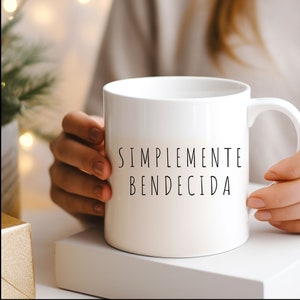 May include: White ceramic mug with the text "SIMPLEMENTE BENDECIDA" in black. The mug is held by a person. The mug is on a white surface. The background has a blurred effect with a Christmas tree and lights.