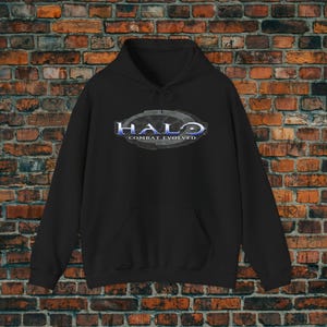 Könnte beinhalten: Schwarzer Kapuzenpullover mit dem Text "HALO COMBAT EVOLVED" in Blau und Silber. Der Pullover hat eine Vordertasche und eine Kapuze mit Kordelzug. Der Hintergrund ist eine Backsteinmauer.