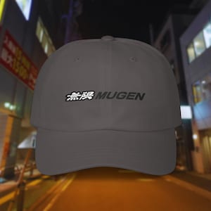 May include: A gray baseball cap with the word "MUGEN" in black and Japanese characters embroidered on the front. The cap has a curved brim and a classic six-panel design. The background is a blurry street scene.