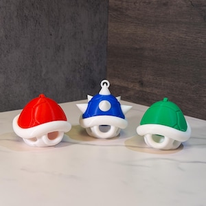 May include: Three colorful, 3D-printed turtle shells in red, blue, and green. Each shell has a white base with a rounded opening and a matching white trim. The blue shell has white spikes.