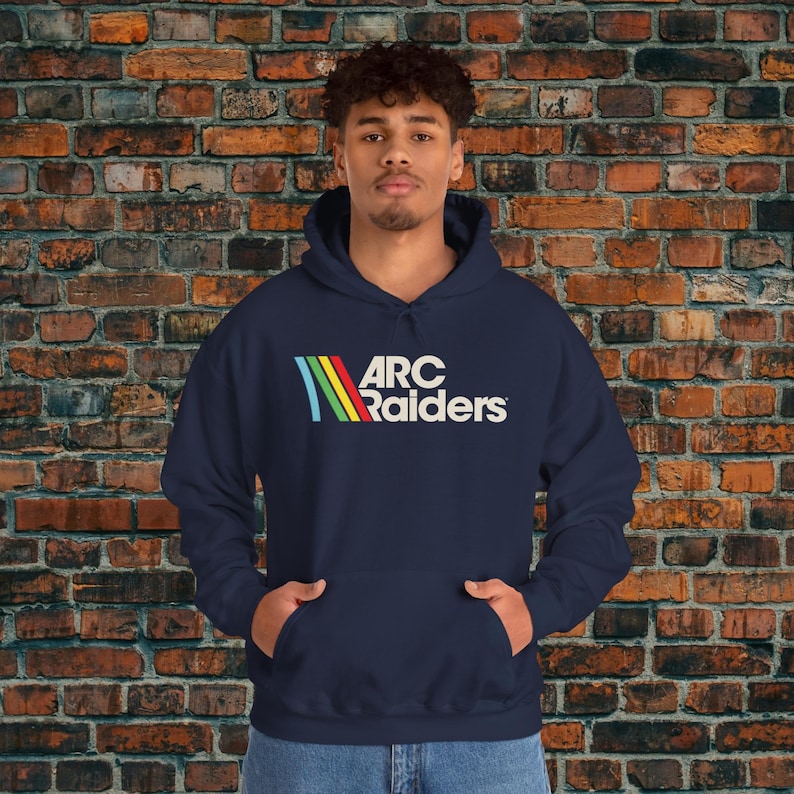May include: Navy blue hooded sweatshirt with the text "ARC Raiders" in white, with a colorful stripe design. The sweatshirt has a front pocket and a drawstring hood. The model is wearing the sweatshirt in front of a brick wall.