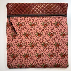 May include: Quilted fabric case with a floral pattern in shades of pink, red, and green. The top section features a brown geometric design. A brown zipper runs across the top, and a beaded pull is attached. The case is designed to hold items.