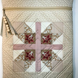 May include: A rectangular quilted pouch featuring a central patchwork block with pink, cream, and floral fabric squares. The exterior is a beige fabric with a fine circular pattern. A beige zipper closure includes a pull with wooden beads and metal accents.