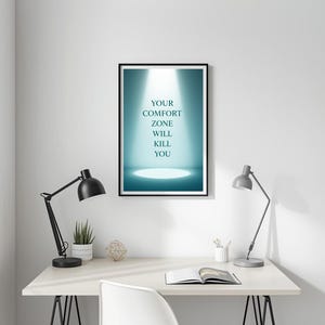 May include: A framed print with the text "YOUR COMFORT ZONE WILL KILL YOU" in white letters against a teal spotlight background. The print is on a white desk with a black desk lamp, a small potted plant, and a geometric decoration.