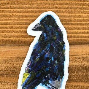 May include: A black crow sticker with a blue and yellow collage design. The crow is standing on a branch with its wings spread out.