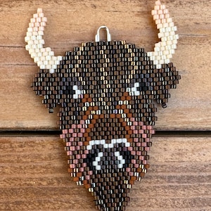 May include: A beaded bull's head ornament with brown, white, and pink beads. The ornament has a loop at the top for hanging.