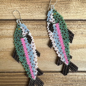 May include: A pair of beaded fish earrings with a green, pink, blue and white colour scheme. The fish are detailed with black beading and have silver hooks.
