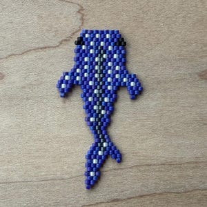 May include: A beaded whale pin in shades of blue, black, and white. The whale is crafted with small beads, creating a textured surface. The pin is set against a wooden background.