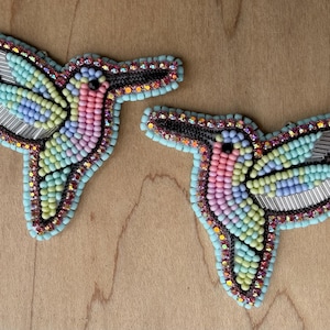 Op de afbeelding: Twee geborduurde kolibrie-broches. Elke broche heeft een kleurrijk ontwerp met roze, blauwe, gele en groene kralen. De vleugels zijn gemaakt van zilverkleurig metaal. De broches zijn omrand met zwarte en turquoise kralen.