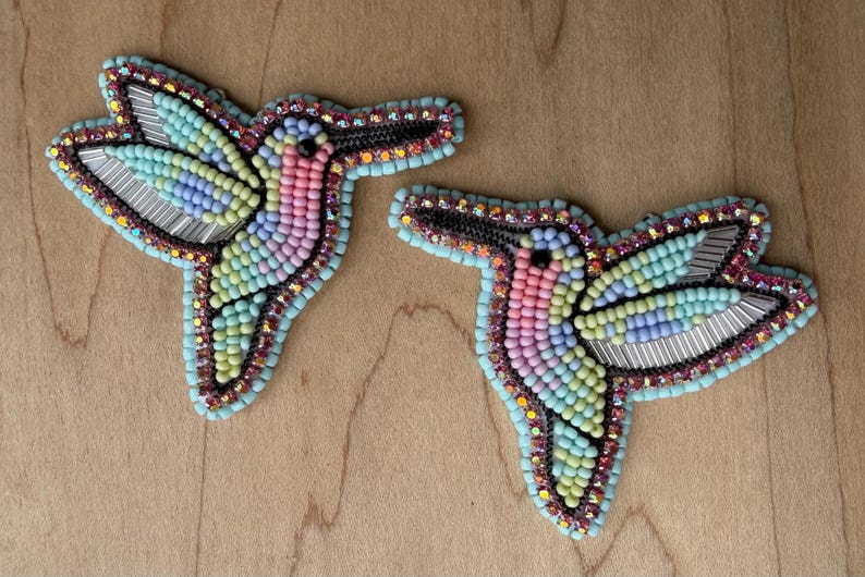 Op de afbeelding: Twee geborduurde kolibrie-broches. Elke broche heeft een kleurrijk ontwerp met roze, blauwe, gele en groene kralen. De vleugels zijn gemaakt van zilverkleurig metaal. De broches zijn omrand met zwarte en turquoise kralen.