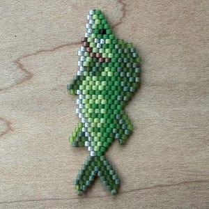 Könnte beinhalten: Ein fischförmiges Ornament aus Perlen in Grün-, Braun- und Silbertönen. Die Perlen sind so angeordnet, dass sie eine strukturierte Oberfläche bilden. Das Ornament liegt auf einer Holzoberfläche.