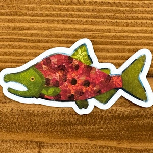 Könnte beinhalten: Ein bunter Lachs-Aufkleber mit einem roten, grünen und braunen Muster. Der Aufkleber befindet sich auf einem hölzernen Hintergrund.