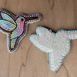 Op de afbeelding: Twee met kralen versierde ornamenten in de vorm van een kolibrie. E&eacute;n is volledig met kralen versierd met een kleurrijk ontwerp, de andere is een witte glitterstof met een lichtblauwe kralenrand. De kolibrie heeft een lichtblauwe kralenrand.