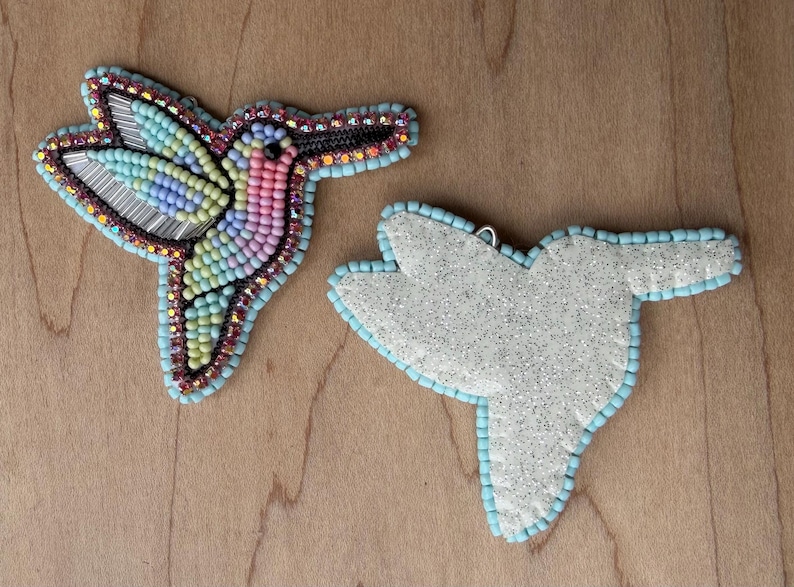 Op de afbeelding: Twee met kralen versierde ornamenten in de vorm van een kolibrie. E&eacute;n is volledig met kralen versierd met een kleurrijk ontwerp, de andere is een witte glitterstof met een lichtblauwe kralenrand. De kolibrie heeft een lichtblauwe kralenrand.