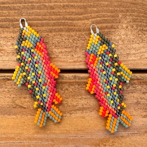 May include: A pair of beaded fish earrings. The earrings are made with small, colorful beads in shades of orange, red, yellow, green, and blue. The fish-shaped earrings have silver-colored ear wires and are displayed on a wooden surface.