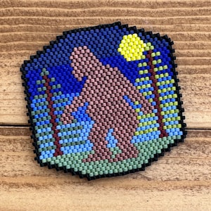 May include: A beaded artwork depicting a brown Bigfoot standing in a forest with a yellow moon in the sky. The background is blue with green and brown trees.