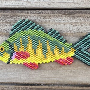 Puede incluir: Un amuleto de pez de cuentas con cuentas verdes, amarillas, rojas y rosas. El pez está hecho con cuentas pequeñas y cuadradas y tiene un diseño detallado.