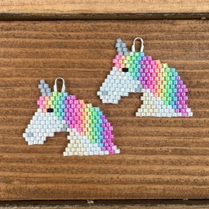 Puede incluir: Dos pendientes de unicornio arcoíris con ganchos plateados. Las cabezas de unicornio están hechas de cuentas blancas e iridiscentes arcoíris.