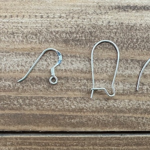 Op de afbeelding: Vier zilverkleurige oorhaakjes in verschillende stijlen worden getoond tegen een houten achtergrond. De haakjes omvatten vis-, herdershaak- en nierdraadontwerpen, die verschillende opties voor het maken van sieraden laten zien.