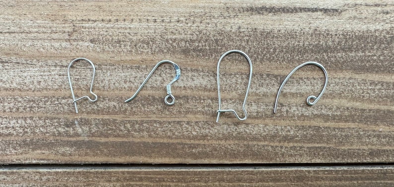 Op de afbeelding: Vier zilverkleurige oorhaakjes in verschillende stijlen worden getoond tegen een houten achtergrond. De haakjes omvatten vis-, herdershaak- en nierdraadontwerpen, die verschillende opties voor het maken van sieraden laten zien.