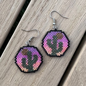 May include: A pair of hexagonal beaded earrings with a pink, green, and brown geometric pattern. The earrings are attached to silver hooks.