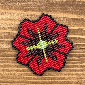 Könnte beinhalten: Eine rote, schwarze und gelbe Perlenblume mit sternförmiger Mitte. Die Blume besteht aus kleinen, quadratischen Perlen und hat eine leicht strukturierte Oberfläche.
